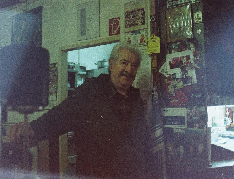 Ein analoges Farbfoto von Aenestis, einem älteren weißhaarigen Mann, der angelehnt an den Tresen in die Kamera lächelt.