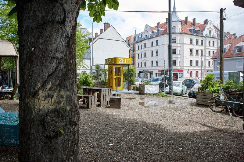 Ein Farbfoto von der ZEITKAPSEL (gelbe Telefonzelle) auf dem Giesinger Grünspitz, einfach Freifläche mit Kies und Bäumen. Im Hintergrund sind Wohnhäuser zu sehen.