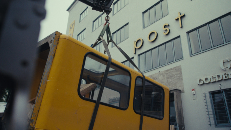 Ein Farbfoto vom Aufbau der Telefonzelle. Zu sehen ist der Transport der Zelle zum Standort vor der TeLa Post, sie schwebt durch die Luft.