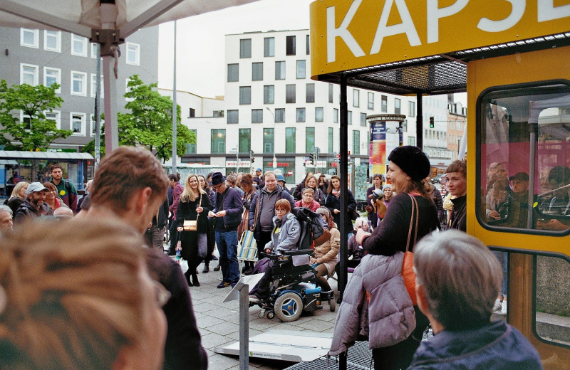Ein analoges Farbfoto von der ZEITKAPSEL (gelbe Telefonzelle mit Dachkonstruktion). Vor ihr stehen viele Menschen, es ist ein Eröffnungsfest.