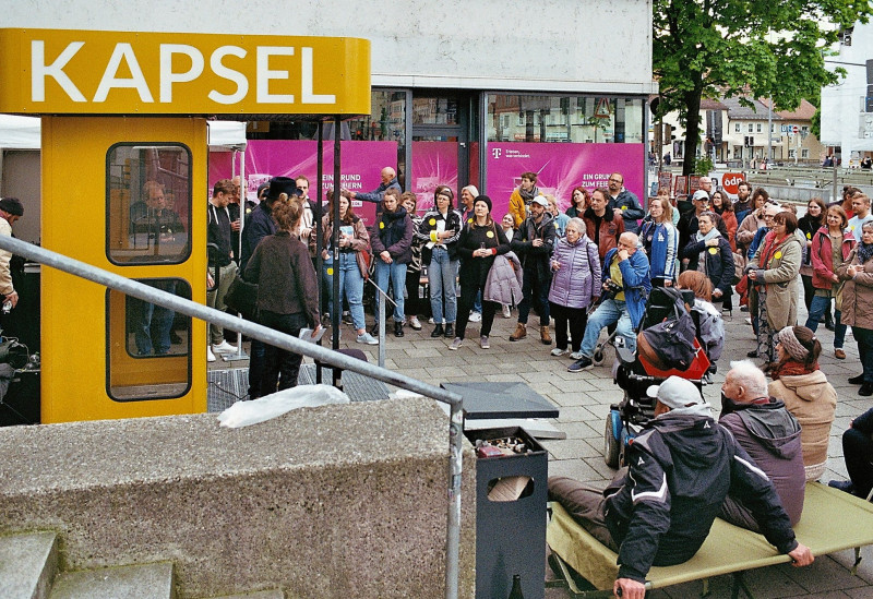 Ein Foto von der ZEITKAPSEL bei der Vernissage im Mai 2023. Es stehen viele unterschiedliche Menschen vor der gelben Telefonzelle.