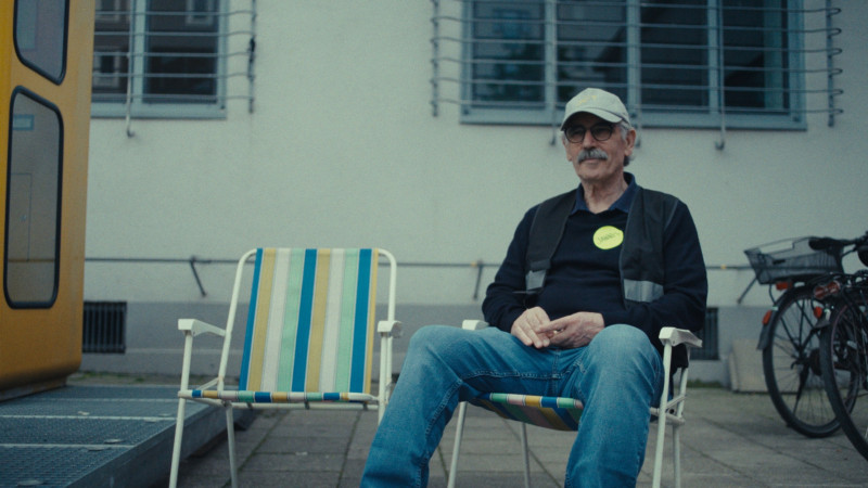 Ein Farbfoto von Jannis bei der Vermittlungsarbeit, er sitzt auf einem von zwei bunt gestreiften Klappstühlen vor der ZEITKAPSEL